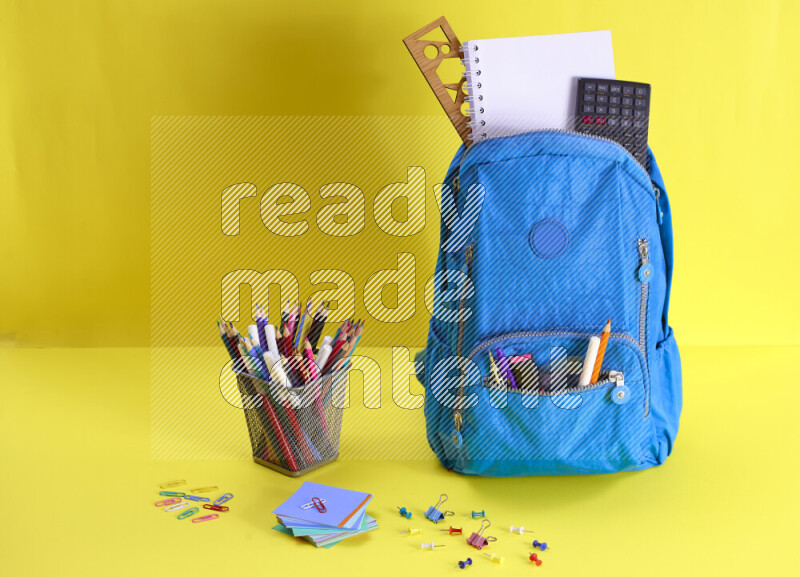 A school bag with assorted school supplies in and beside it on yellow background