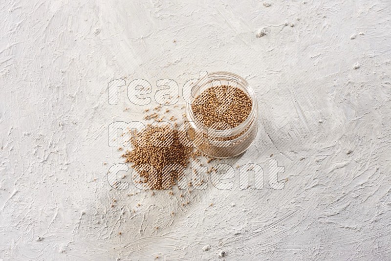 A glass jar full of mustard seeds and more seeds spread on a textured white flooring in different angles