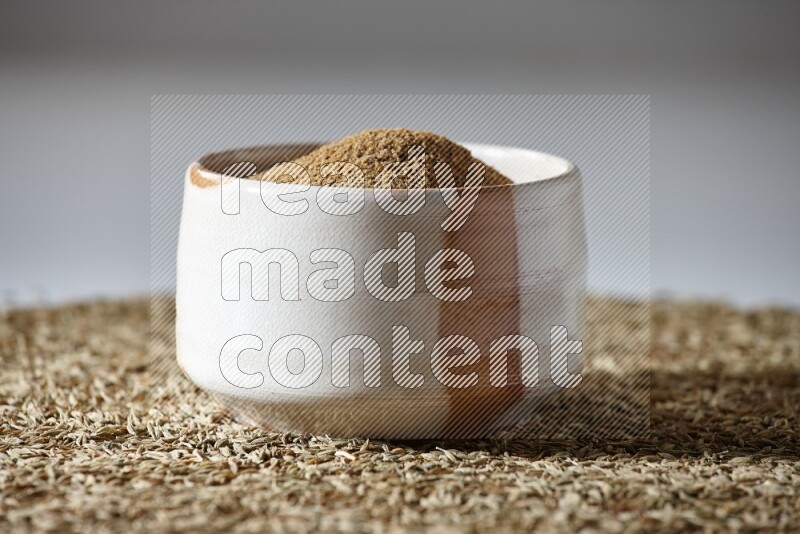 A beige bowl full of cumin powder with cumin seeds beneath it the bowl on a white flooring