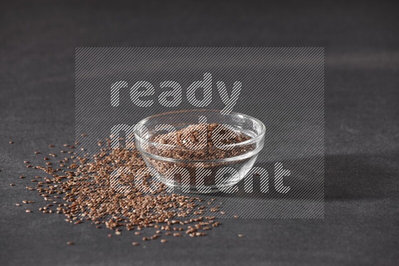 A glass bowl full of flaxseeds surrounded by the seeds on a black flooring