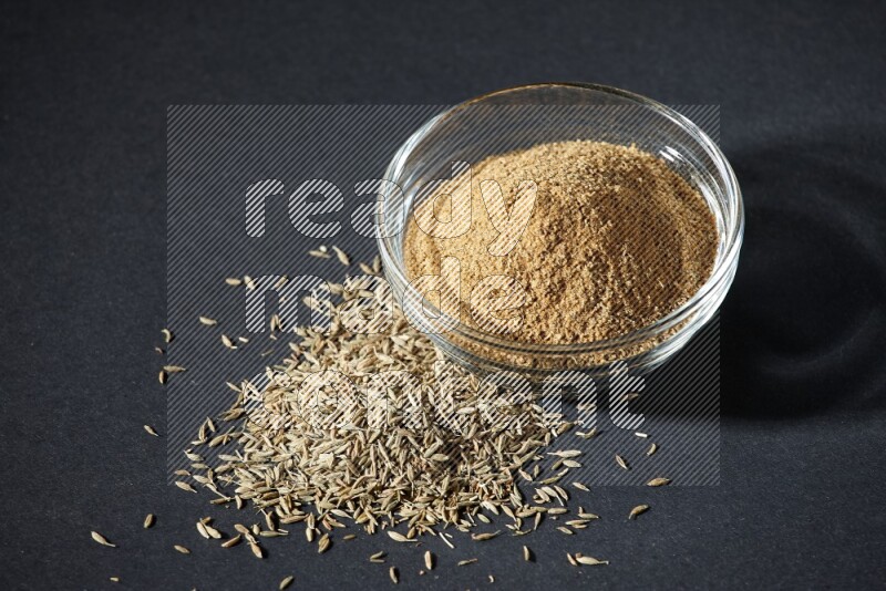 A glass bowl full of cumin powder with cumin seeds beside it on black flooring
