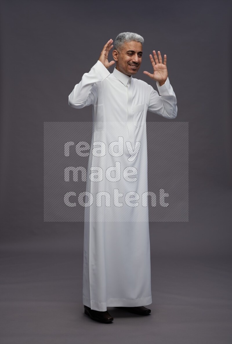 Saudi man wearing thob standing hands behind head on gray background