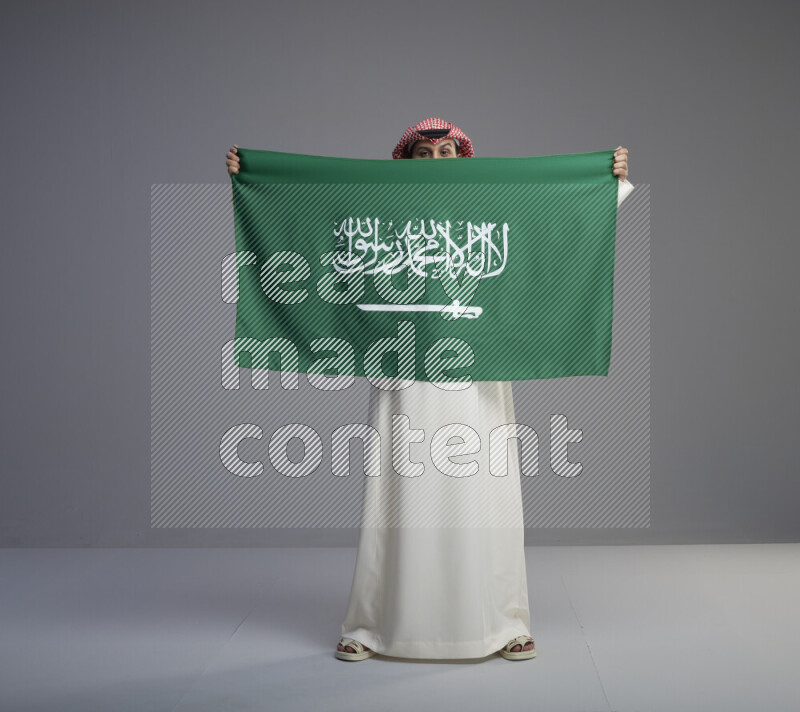 A saudi man standing wearing thob and red shomag holding big saudi flag on gray background