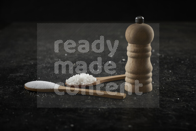 2 wooden spoons filled with fine and coarse salt with wooden grinder beside them on black background