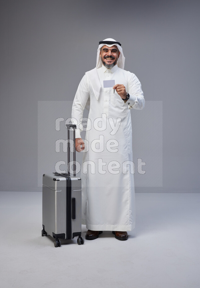 Saudi man wearing Thob and white Shomag standing holding Travel bag and ATM card on Gray background