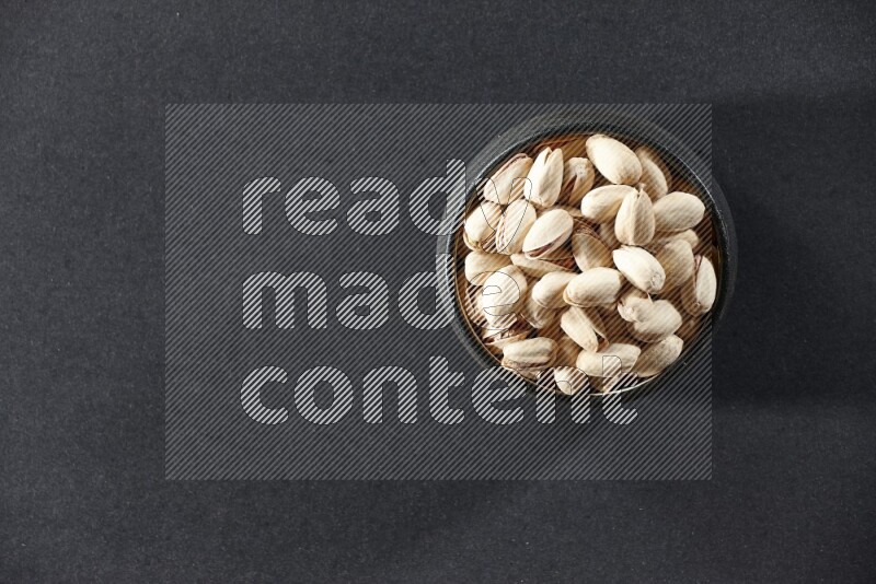 A black pottery bowl full of pistachios on a black background in different angles