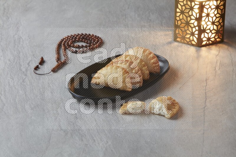 Four fried sambosas in an oval shaped black plate, beside a cut cheese sambosa, a brown misbaha and a golden lantern on a gray background