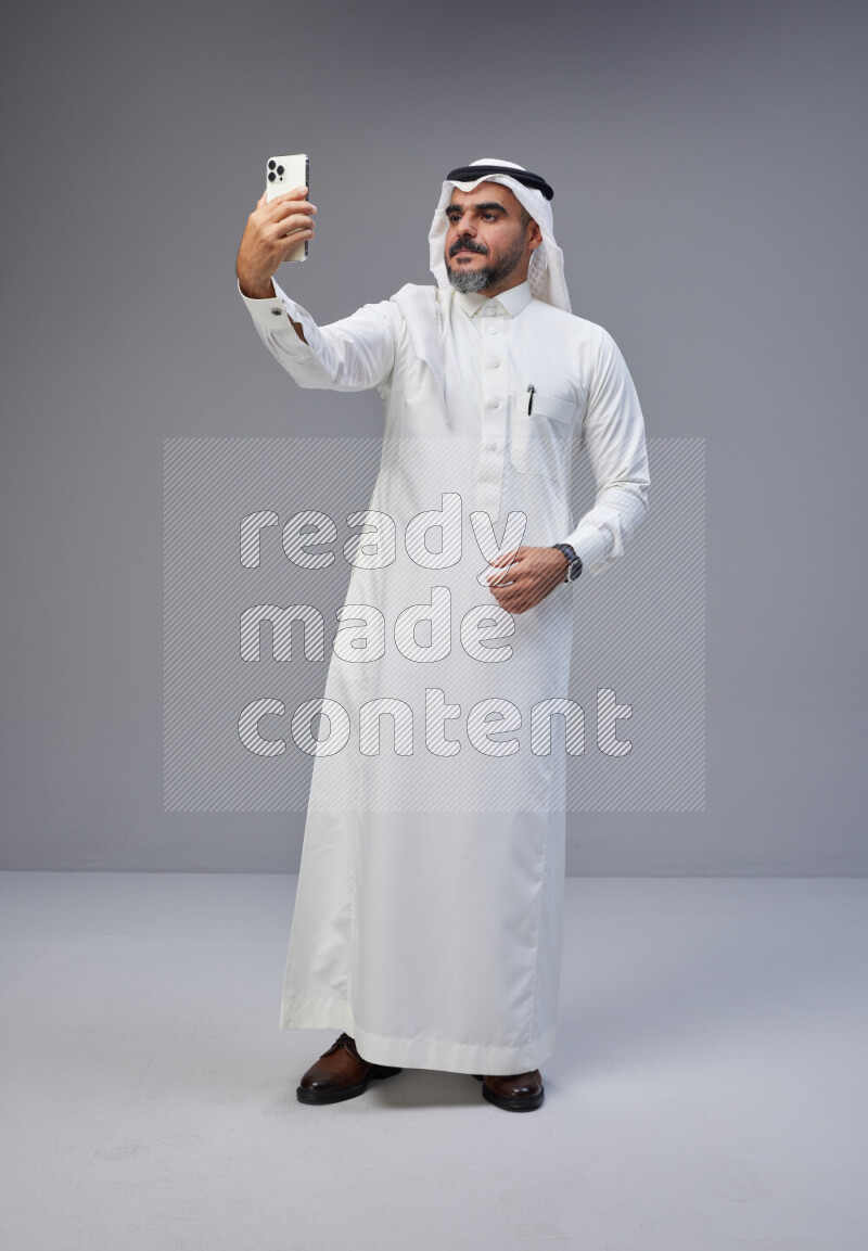 Saudi man Wearing Thob and white Shomag standing taking selfie on Gray background