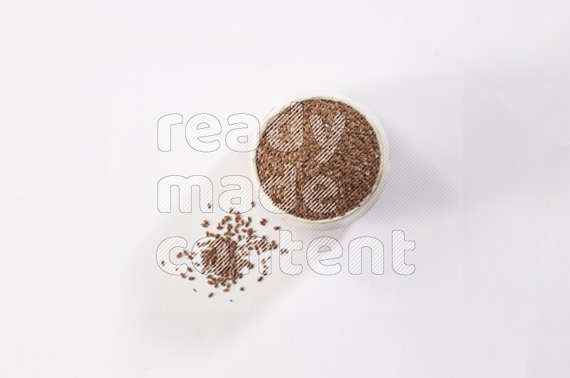 A pottery beige bowl full of flax seeds and more seeds spread on a white flooring