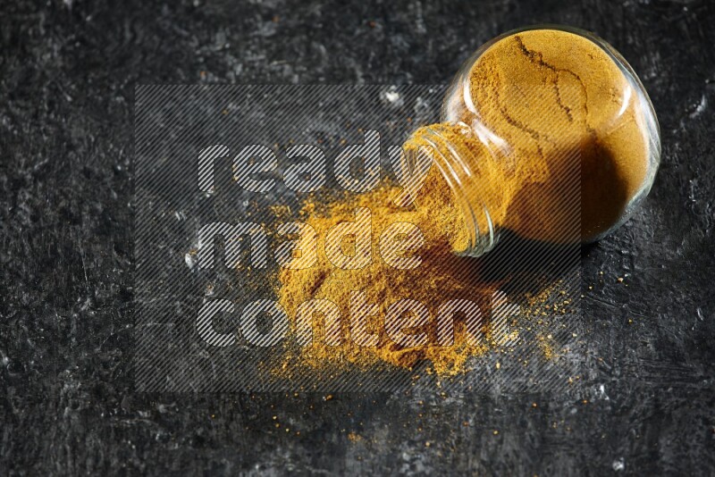 A flipped glass spice jar full of turmeric powder and powder spilled out of it on textured black flooring