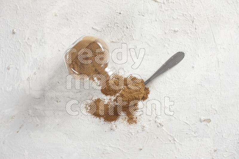 Flipped herbs glass jar full of cinnamon powder with a metal spoon full of powder on a textured white background