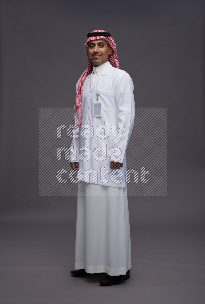 Saudi man wearing thob with lab coat and shomag with pocket employee badge standing interacting with the camera on gray background