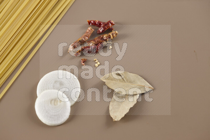 Raw pasta with different ingredients such as cherry tomatoes, garlic, onions, red chilis, black pepper, white pepper, bay laurel leaves, rosemary and cardamom on beige background