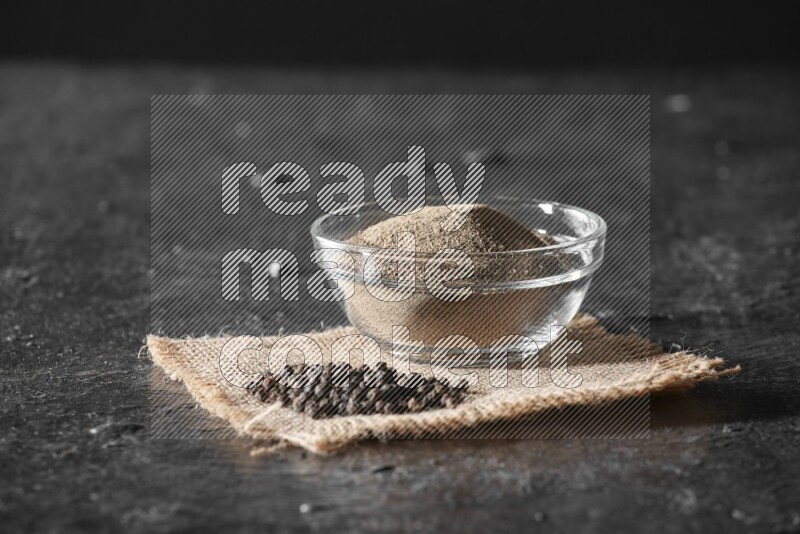 A glass bowl full of black pepper powder and black pepper beads on burlap fabric on textured black flooring