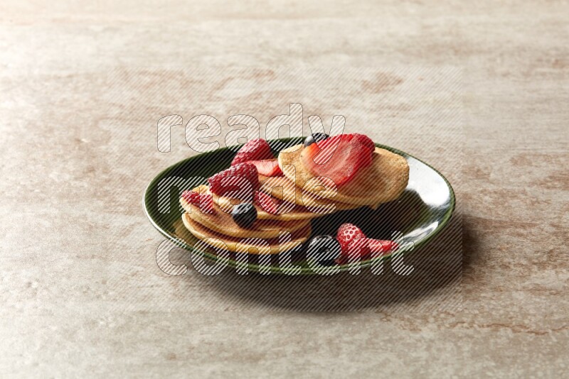 Five stacked mixed berries mini pancakes in a green plate on beige background
