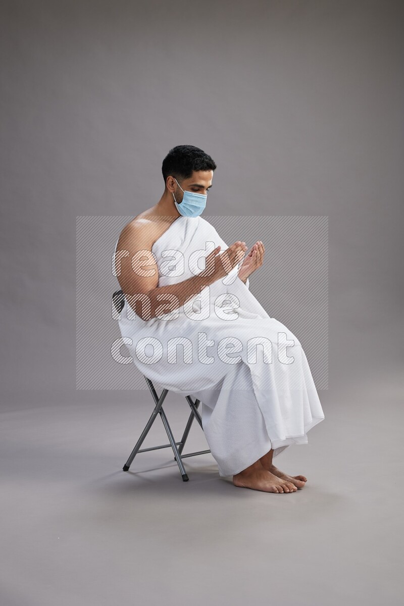A man wearing Ehram with face mask sitting on chair dua'a on gray background