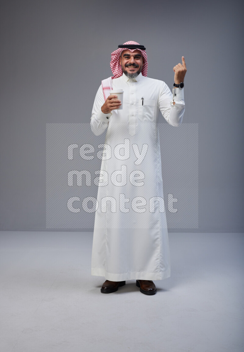 Saudi man Wearing Thob and red Shomag standing holding paper cup on Gray background