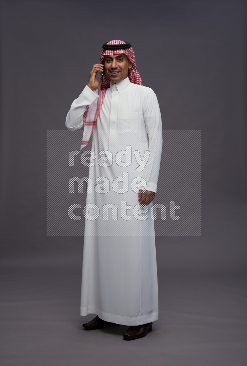 Saudi man wearing thob and shomag standing talking on phone on gray background