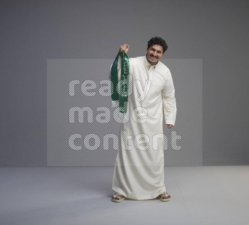 A Saudi man standing wearing thob holding Saudi flag scarf on gray background