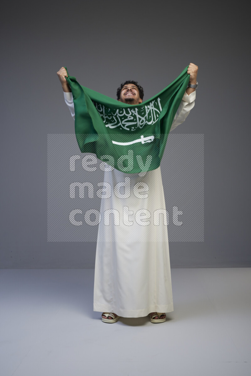 A saudi man standing wearing thob holding big saudi flag on gray background