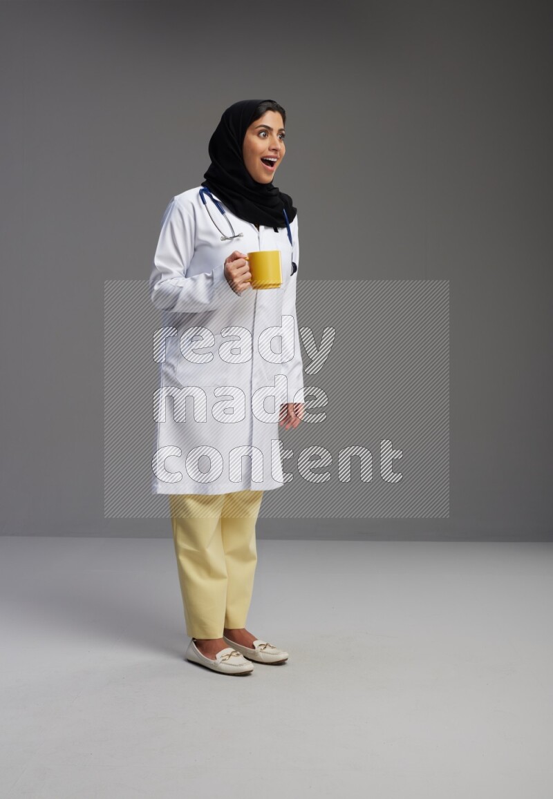 Saudi woman wearing lab coat with stethoscope standing holding mug on Gray background