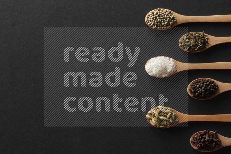 wooden spoons filled with white peppers, cloves, cardamom, salt, black peppers and basil on black flooring