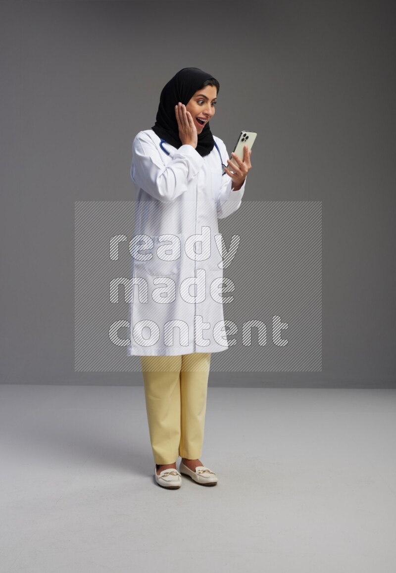 Saudi woman wearing lab coat with stethoscope standing texting on phone on Gray background
