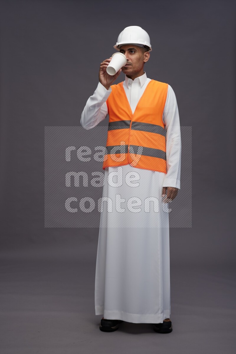 Saudi man wearing thob with engineer vest standing holding paper cup on gray background