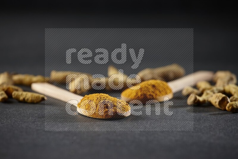 2 wooden spoons full of turmeric powder with dried turmeric fingers on black flooring