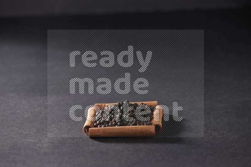 أربعة من أعواد القرفة مكونة شكل مربع بداخلها حبوب الفلفل الأسود علي خلفية سوداء