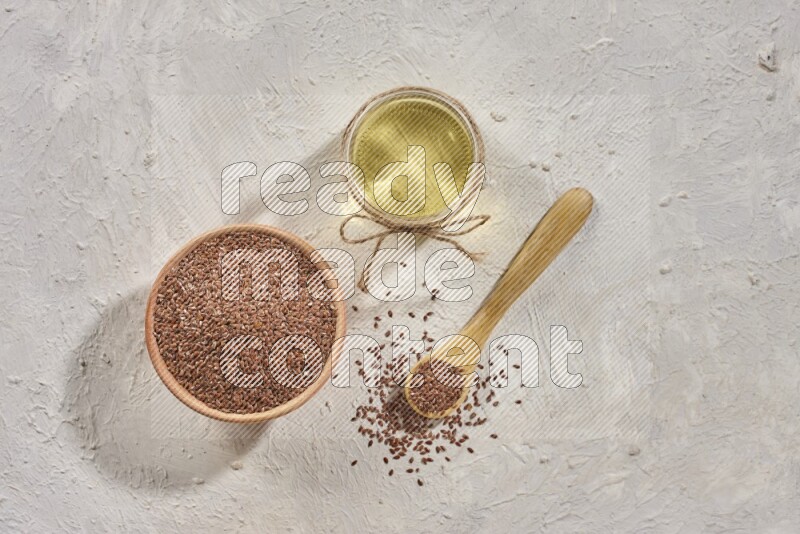 A wooden bowl and spoon full of flax seeds and a glass jar of flaxseeds oil on a textured white flooring