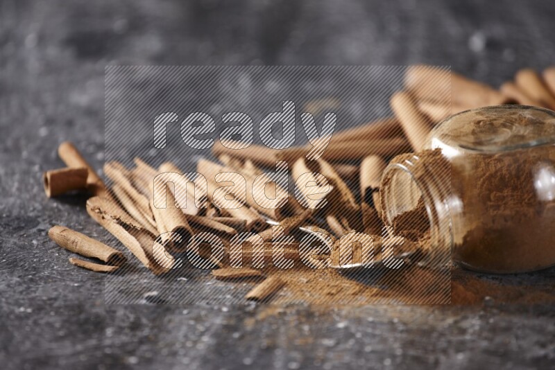 Herbal glass jar full cinnamon powder flipped and a metal spoon full of powder surrounded by cinnamon sticks on textured black background in different angles