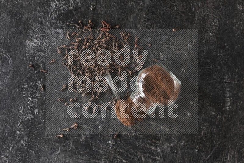 A flipped glass spice jar and a metal spoon full of cloves powder with cloves spread on textured black flooring