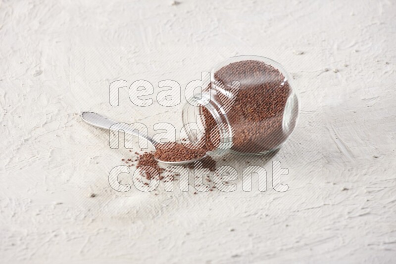 A glass spice jar and a metal spoon full of garden cress seeds and jar is flipped with fallen seeds on a textured white flooring