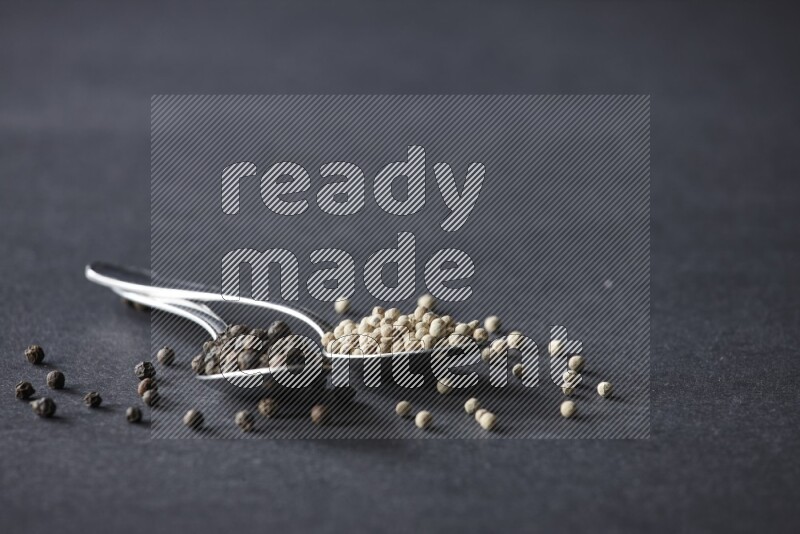 2 metal spoons full of black and white pepper beads with spreaded beads on black flooring