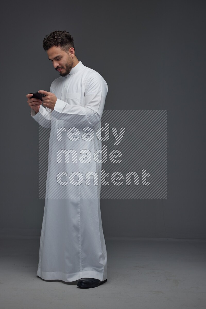 A Saudi man wearing Thobe and playing using his phone horizontally playing video games while standing and making different poses eye level on a gray background