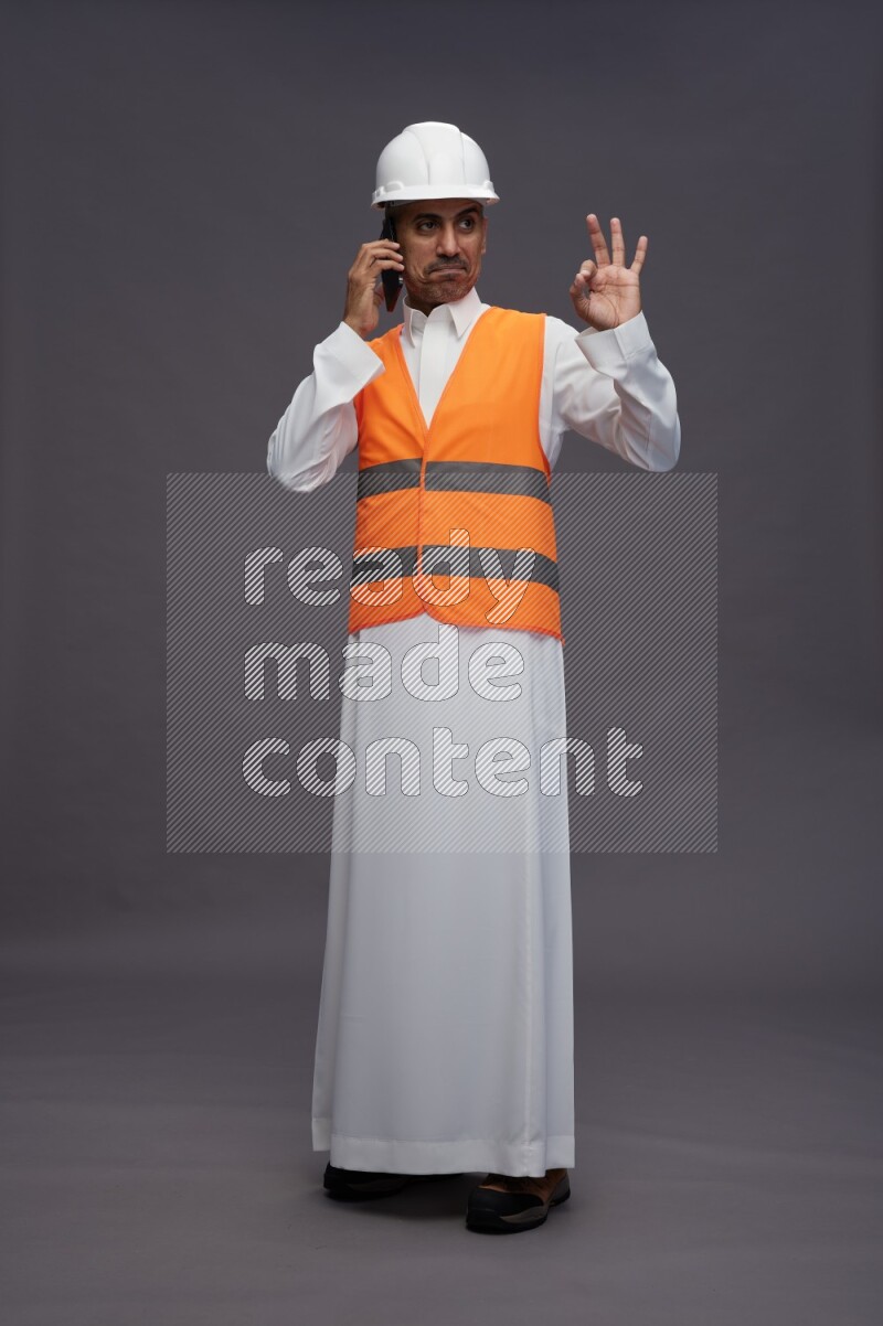 Saudi man wearing thob with engineer vest standing talking on phone on gray background