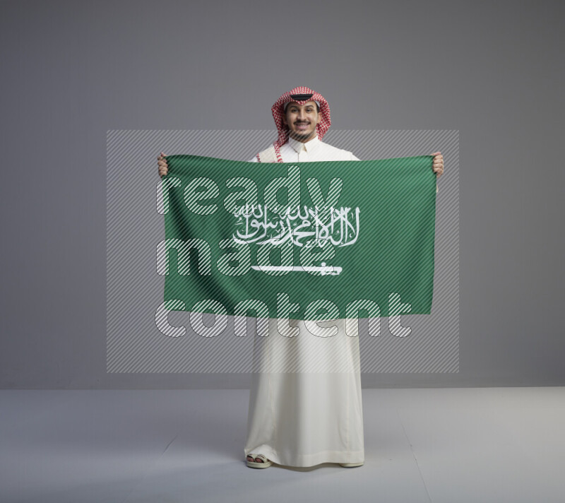 A saudi man standing wearing thob and red shomag holding big saudi flag on gray background