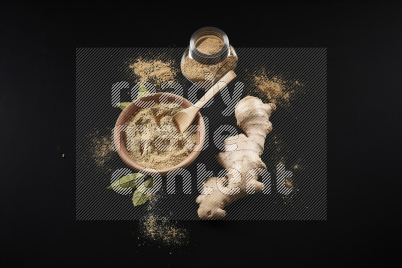 A wooden bowl full of ground ginger powder with a glass jar beside it and fresh ginger on black background