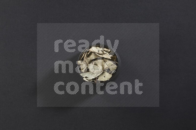 A black pottery bowl full of dried bay leaves on black flooring