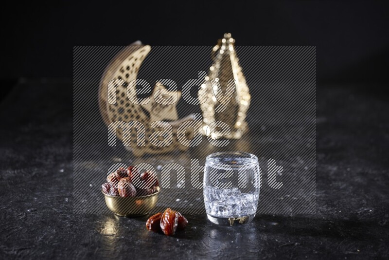 Dates in a metal bowl with water beside golden lanterns in a dark setup