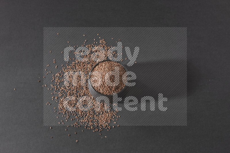 A black pottery bowl full of flaxseeds surrounded by the seeds on a black flooring