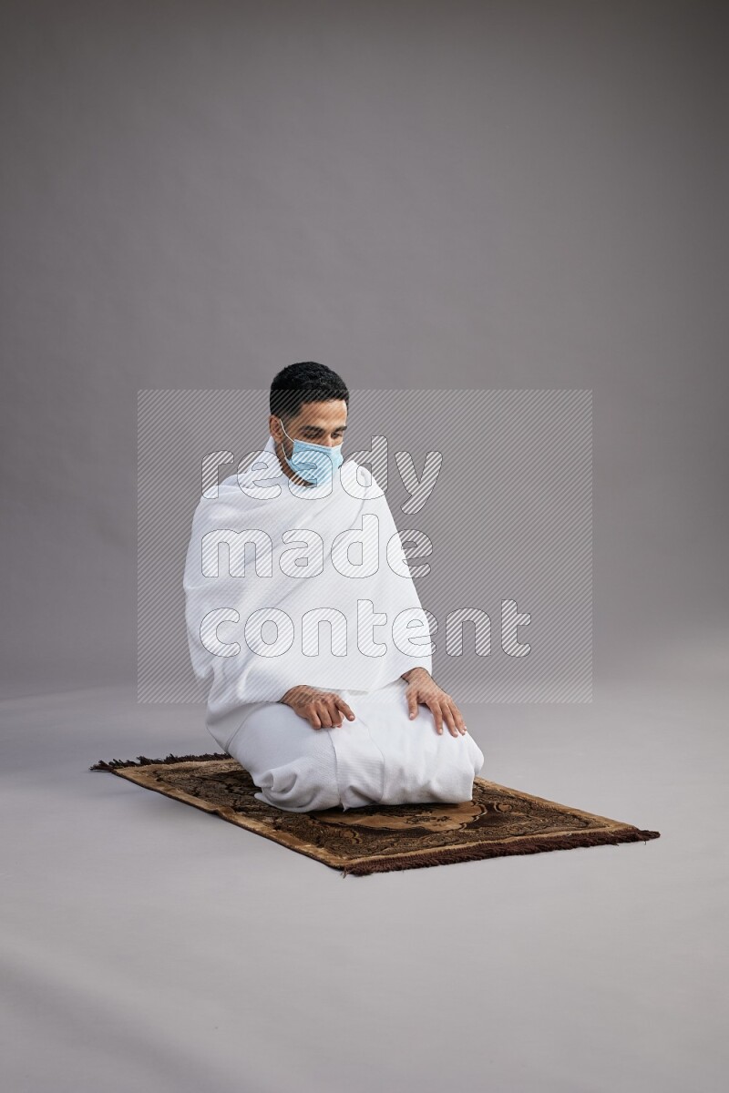 A man wearing Ehram with face mask sitting on floor performing dua'a on gray background