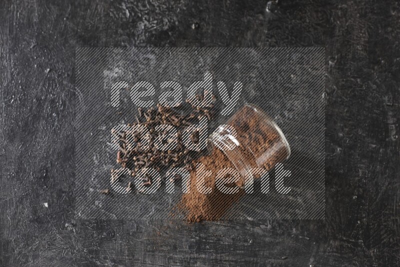 A flipped glass jar full of cloves powder with cloves spread on a textured black flooring