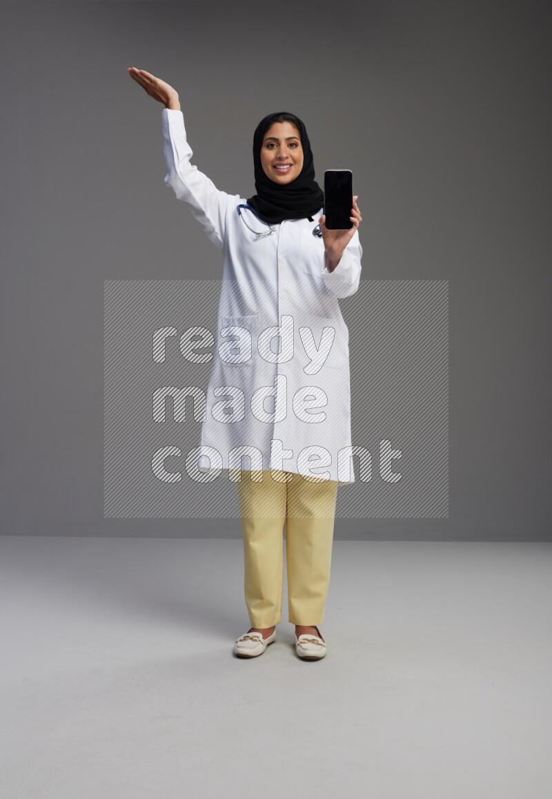 Saudi woman wearing lab coat with stethoscope standing showing phone to camera with sign in the back on Gray background
