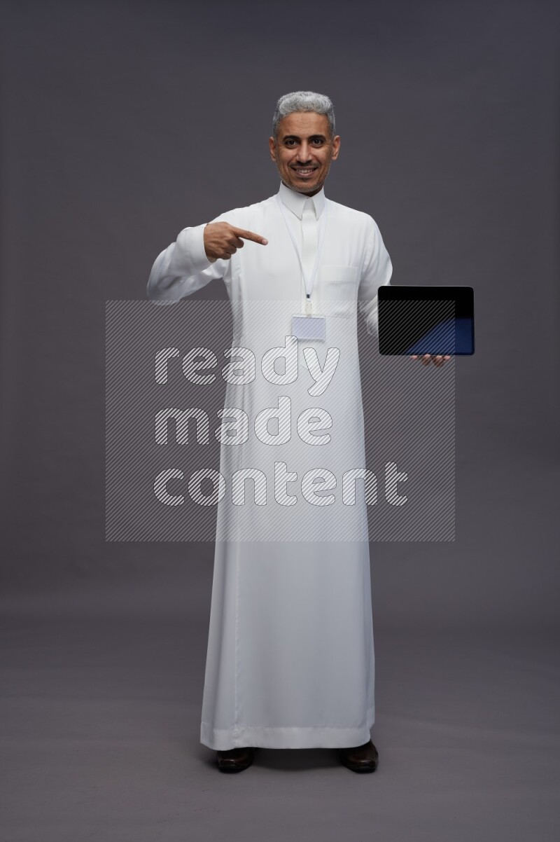 Saudi man wearing thob with neck strap employee badge standing showing tablet to camera on gray background
