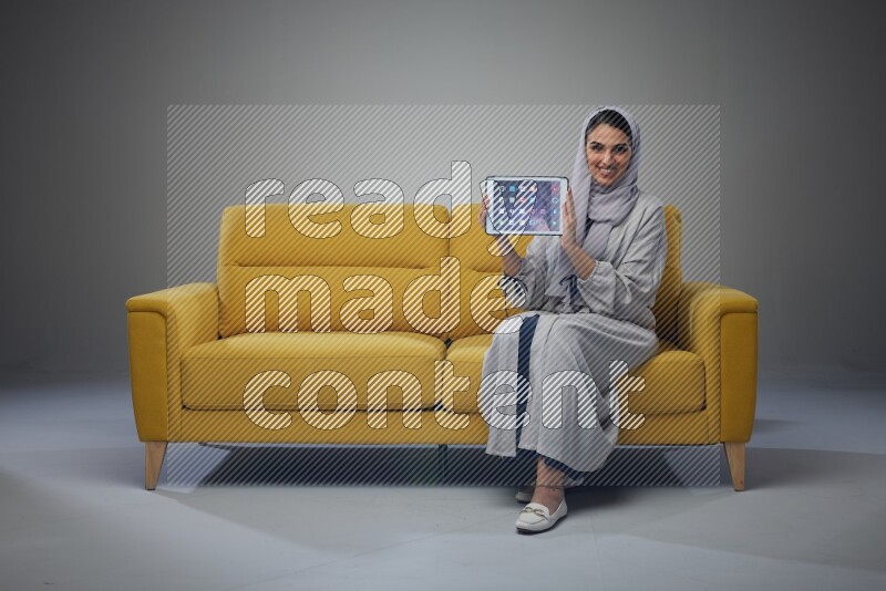 A Saudi female wearing a light gray Abaya and head scarf sitting on a yellow sofa and showing her tablet's screen while wearing headphones eye level on a grey background