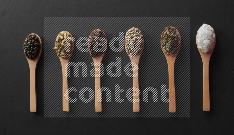 wooden spoons filled with white peppers, cloves, cardamom, salt, black peppers and basil on black flooring