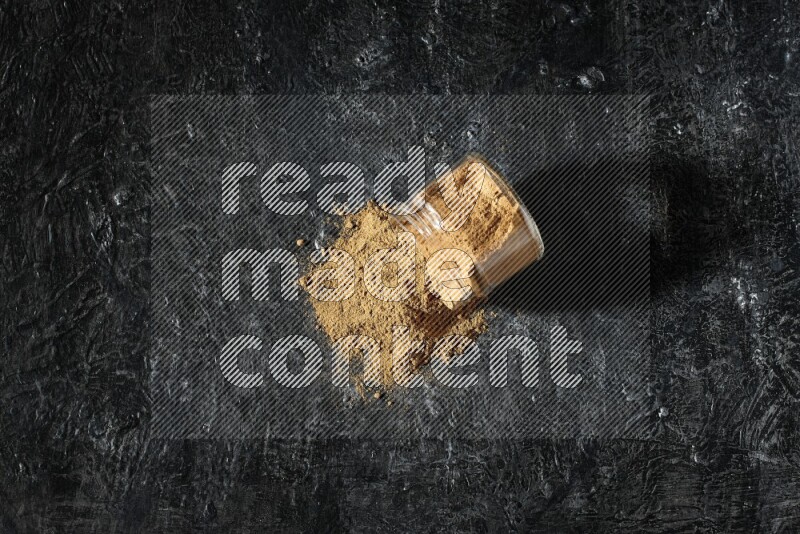 A flipped glass jar full of allspice powder and powder spilled out of it on a textured black flooring
