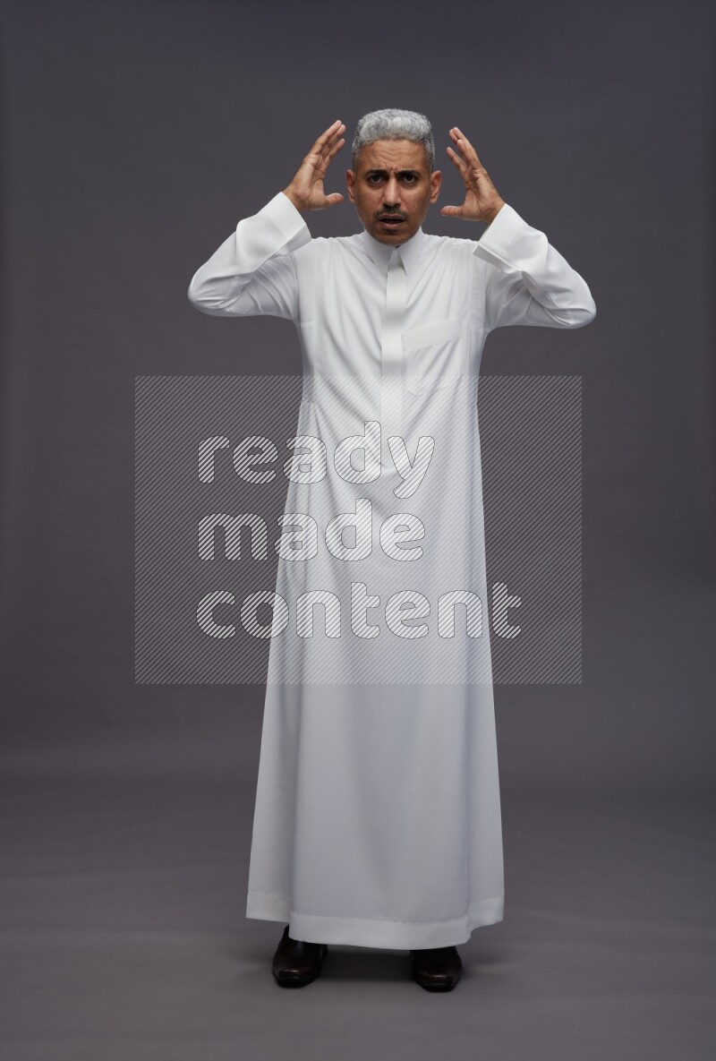 Saudi man wearing thob standing hands behind head on gray background